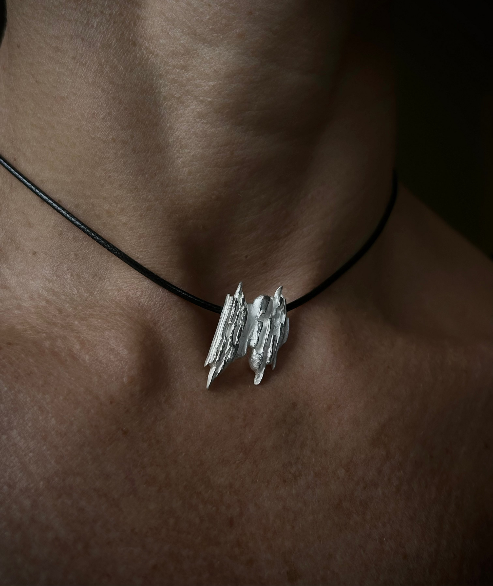 Silver pendant on a black cord necklace worn by a person with a dark background